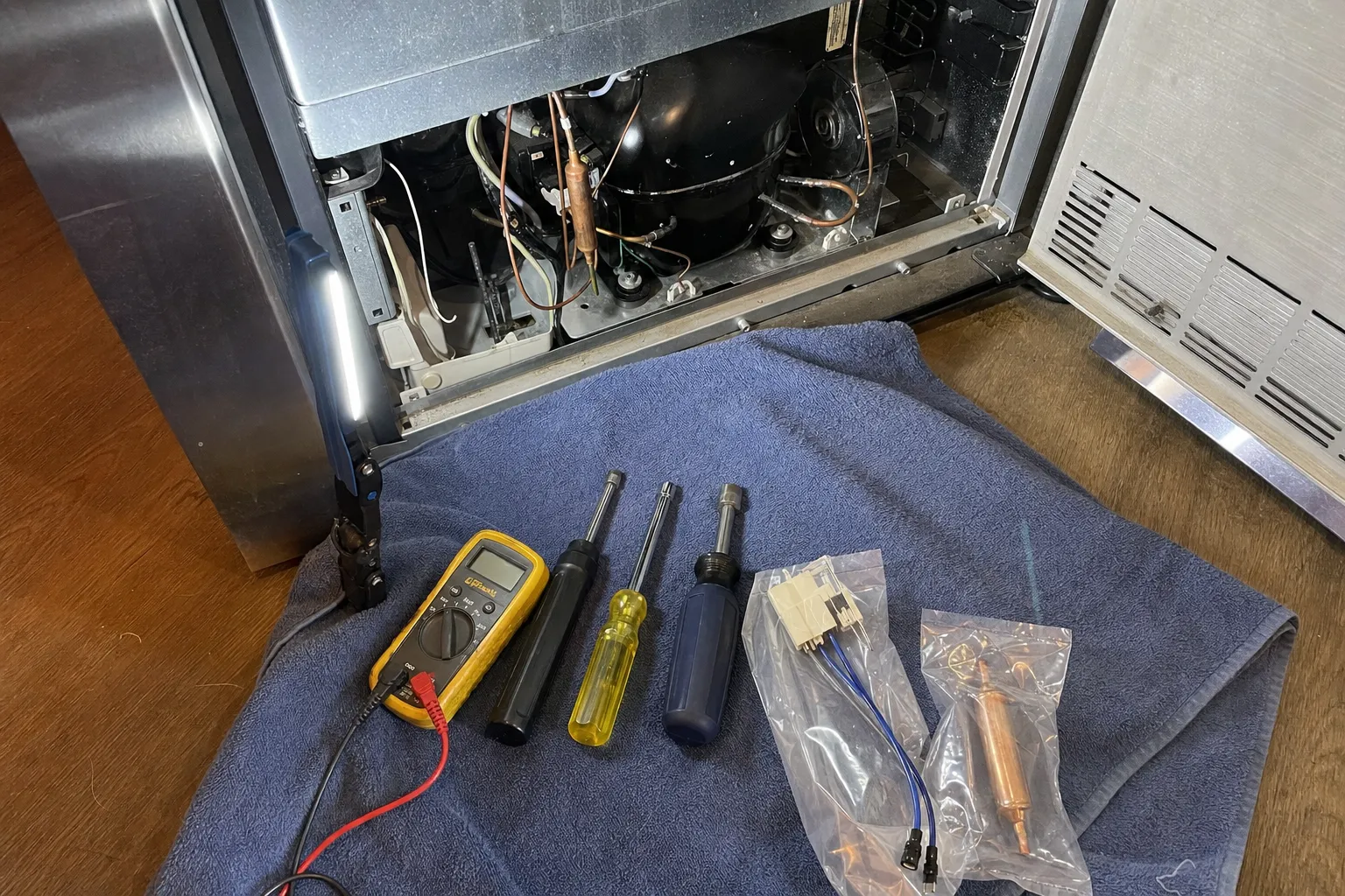 Multimeter, nut drivers and bagged replacement relay and filter-drier laid out beside an open compressor bay