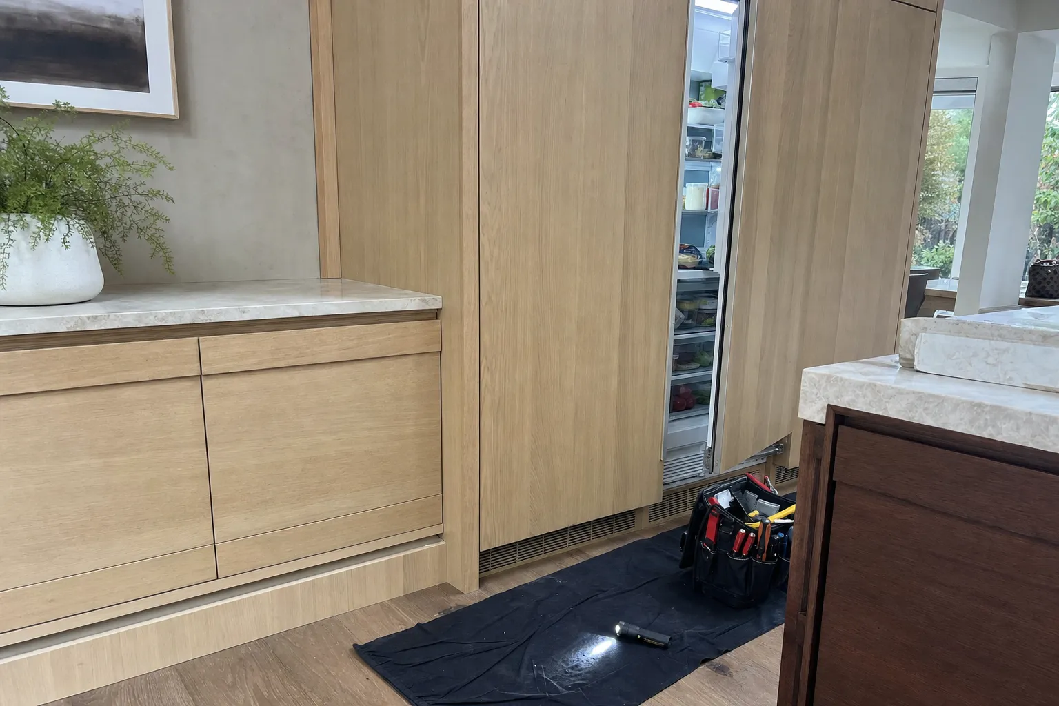 Oak-panelled integrated refrigerator column standing ajar in a run of flush cabinetry, tool bag and flashlight on a mat