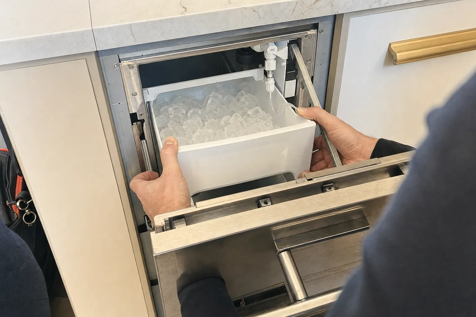 Ice bin of clear cubes lifted from an undercounter ice drawer, the white fill fitting visible above it